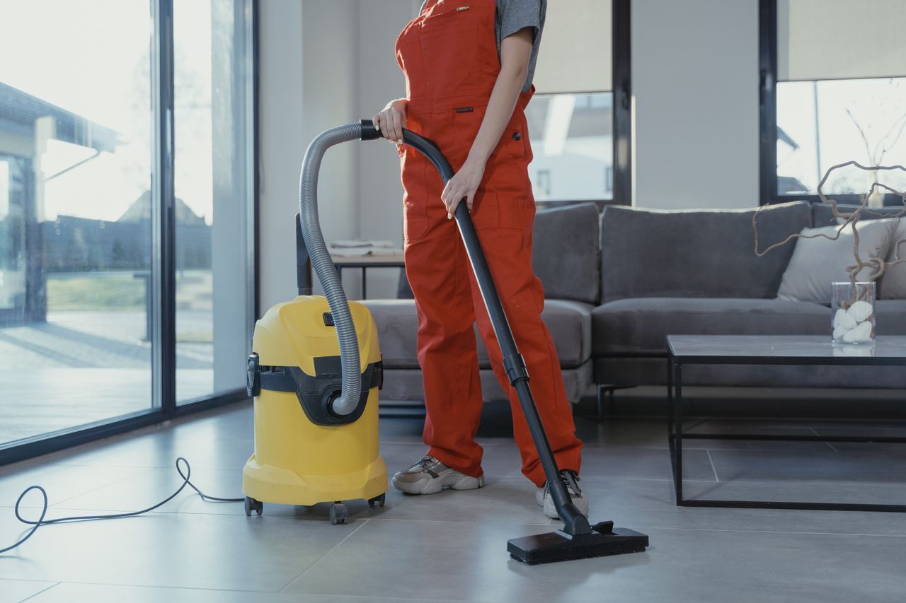 services-03 Un nettoyeur professionnel en combinaison rouge aspirant un salon moderne avec un aspirateur jaune.